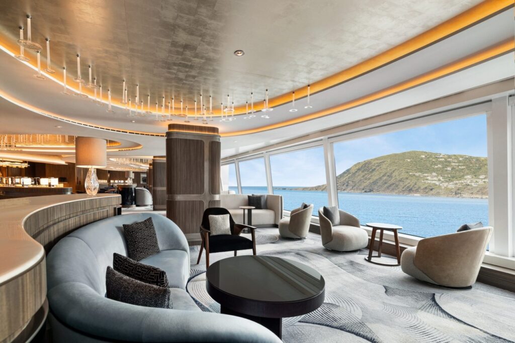 Elegante lounge panorámico de un crucero con sillones modernos y grandes ventanales con vista al mar y a la costa.