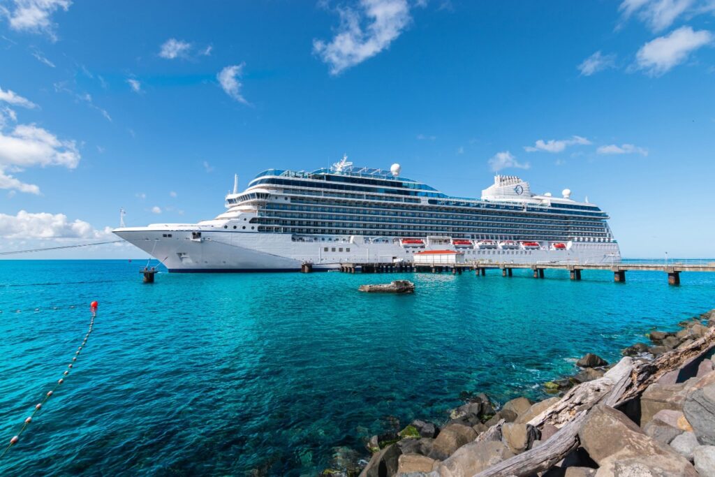Gran crucero blanco atracado en un muelle sobre aguas turquesa bajo un cielo azul con algunas nubes.