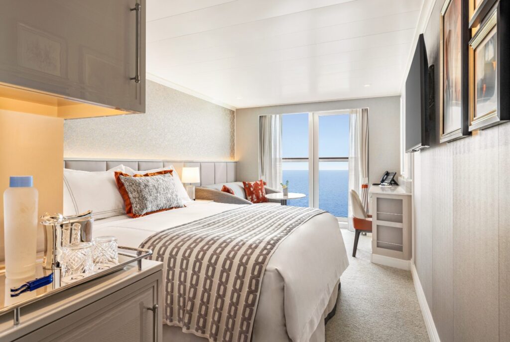 Cabina elegante de crucero con cama grande, decoración moderna y puertas de vidrio que dan a una terraza con vista directa al océano.